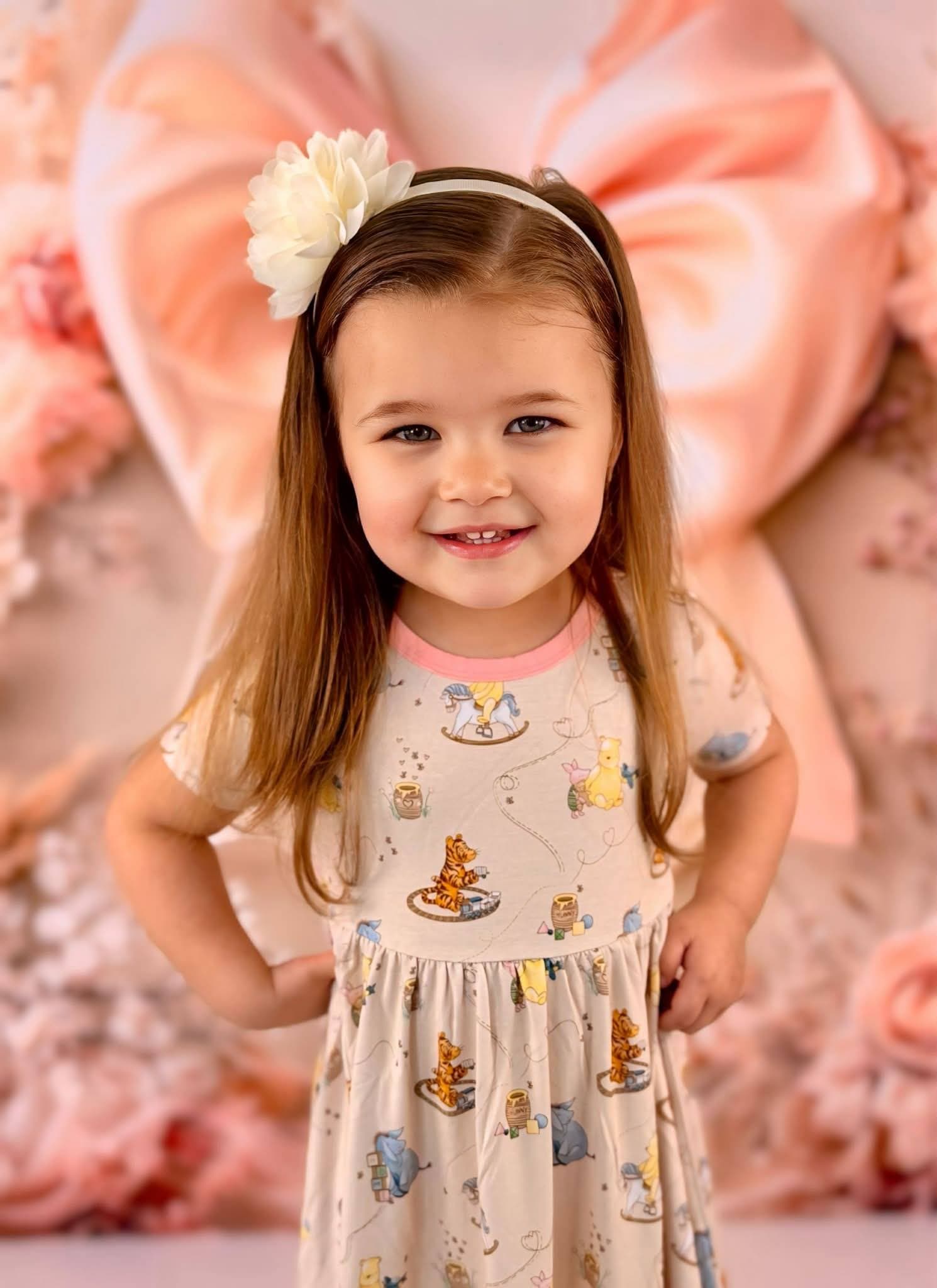 Young girl in a Winnie the Pooh bamboo twirl dress with a floral headband against a pink floral background